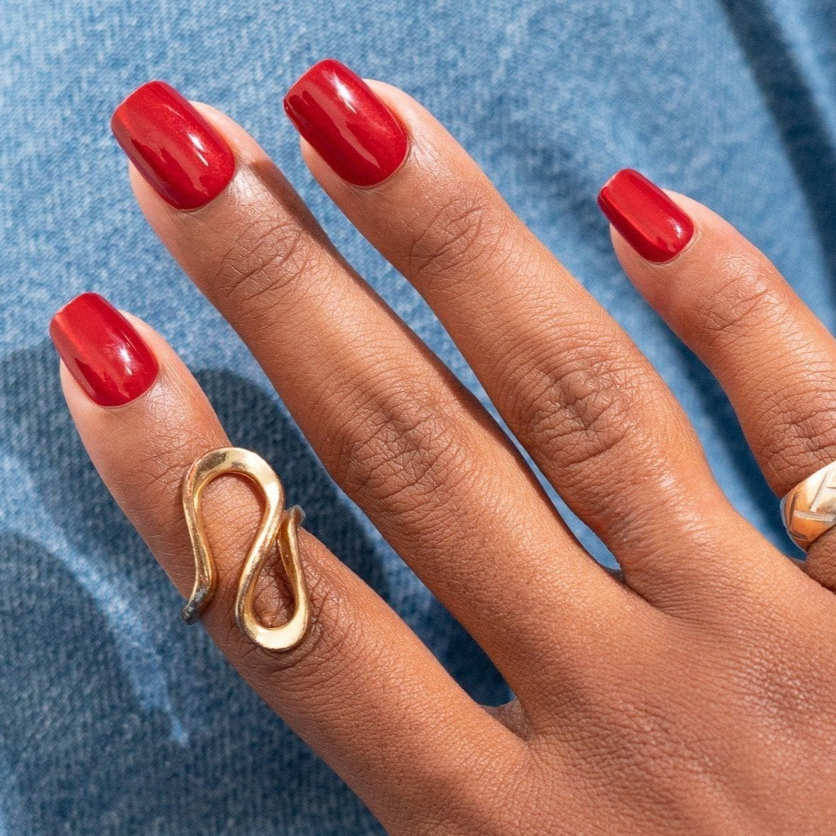 A person wearing ruby red press-on nails and a gold-colored ring, with jeans in the background.